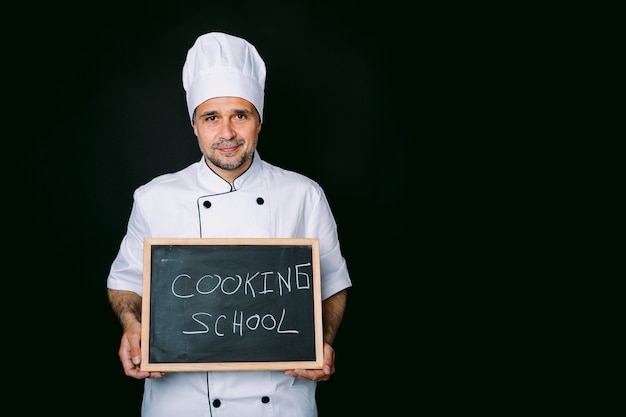 Cook, gekleidet in einen weißen Hut und eine Jacke, hält eine Tafel mit der Aufschrift: Kochschule auf schwarzem Hintergrund. Restaurant, Essen und Kochschulkonzept.