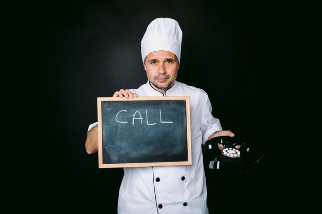 Cook in einer weißen Jacke und Hut hält eine Tafel mit der Aufschrift: "Anruf" und ein Vintage-Telefon auf schwarzem Hintergrund. Restaurant- und Take-away-Konzept.