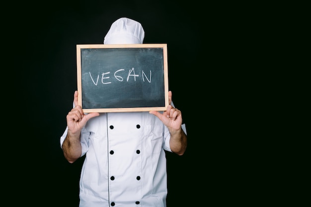 Cook in einer weißen Jacke und Hut hält eine Tafel mit der Aufschrift: "Vegan" auf schwarzem Hintergrund. Veganes Essen und Restaurantkonzept.