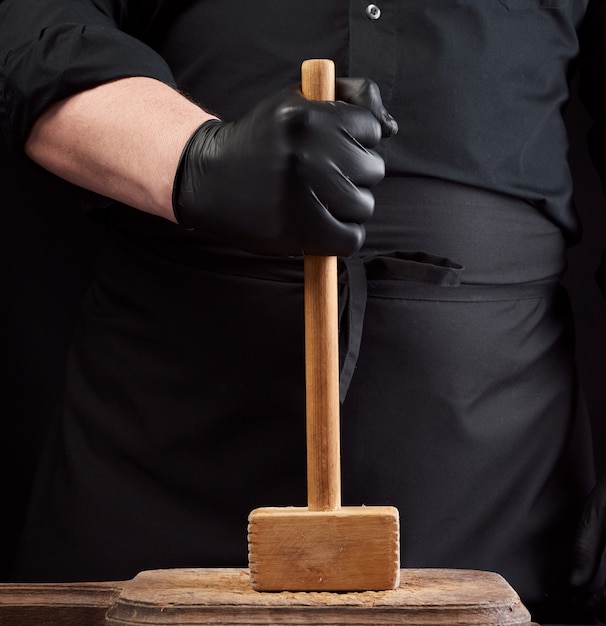 Cook en uniforme negro tiene un martillo de madera para batir carne en una tabla de cortar vintage