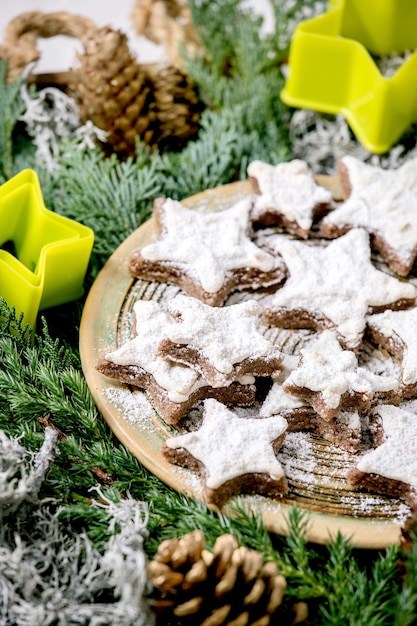 Cookies caseiros de amêndoa de cacau em forma de estrela com esmalte branco e açúcar de confeiteiro. Na placa de cerâmica com cortadores de biscoitos de estrelas de Natal em ramos de thuja. Fechar-se