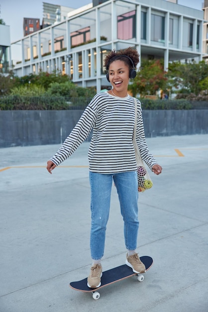 Cool adolescente monta patineta contra el paisaje urbano usa jeans a rayas y zapatillas de deporte escucha la pista de audio favorita se ve con gusto en la distancia le gusta andar en patineta Concepto de cultura juvenil