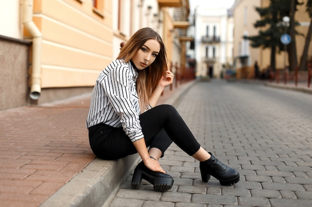 Cool bastante joven en una elegante blusa a rayas en blanco y negro en jeans de moda en zapatos de cuero negro con una nariz perforada descansando sentado al aire libre en la ciudad.