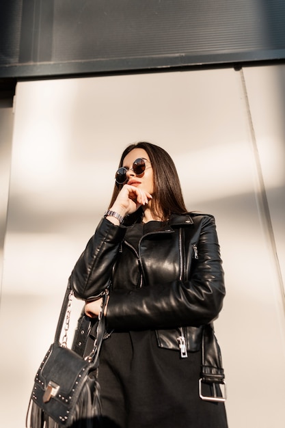 Cool elegante hermosa mujer hipster en ropa de moda rock negro con chaqueta de cuero y gafas de sol redondas vintage cerca de la pared en la ciudad