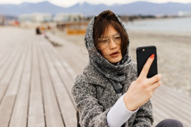 Cool elegante joven morena con gafas hace selfie al aire libre, sopla el viento