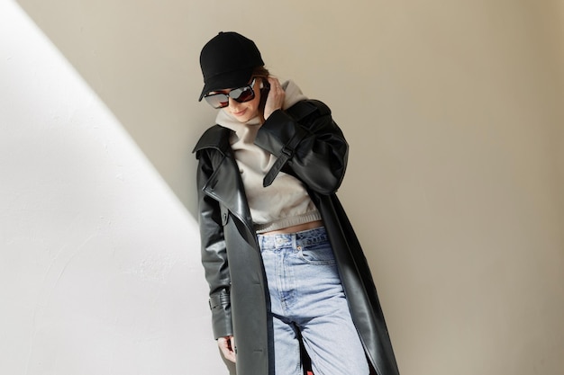 Cool fashion street hermosa mujer hipster con gafas de sol de moda y gorra negra en ropa urbana casual con abrigo con capucha y jeans cerca de una pared