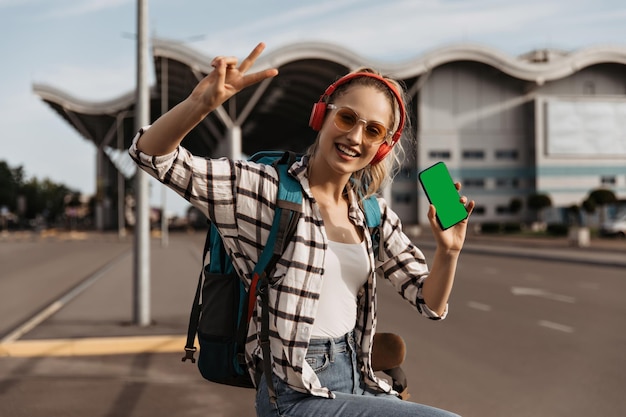 Cool girl en camisa a cuadros y gafas de sol muestra vsign sostiene el teléfono y escucha música en los auriculares Retrato de turista con mochila afuera