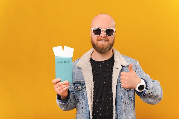 Cool joven calvo barbudo con gafas de sol con pasaporte y pulgar hacia arriba