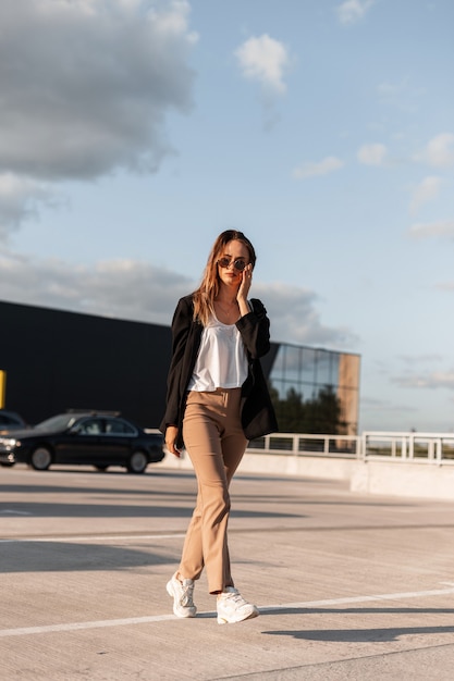 Cool joven con elegante chaqueta negra en pantalones vintage con gafas de sol de moda en la parte superior camina sobre asfalto en el cielo azul de fondo en un día soleado de verano brillante. Viaje de chica de moda en el aparcamiento de la ciudad.