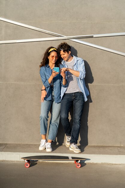 Cool pareja joven mirando en la pantalla del teléfono inteligente al aire libre abrazo stand en longboard skate sonrisa feliz