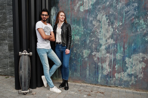 Cool pareja multirracial posando contra la pared con longboard