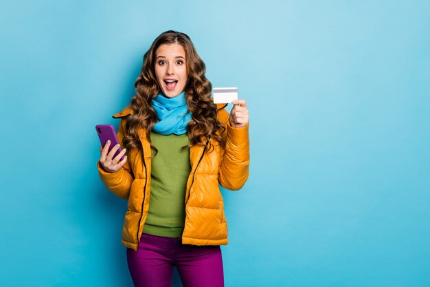 Coole Art zu kaufen. Foto der hübschen Dame mit Telefon, das Online-Zahlung macht, verwendet Plastikkreditkarte, die gelbe Mantelschalhose Pullover isolierte blaue Farbwand trägt