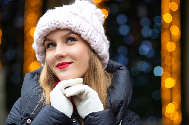 Coole blonde Frau mit rotem Lippenstift mit Strickmütze posiert auf der Straße über einem Girlandenhintergrund mit Bokeh