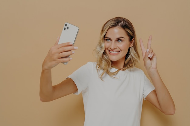 Coole fröhliche Frau mit blonden Haaren mit Smartphone in der Hand, die im Studio vor nacktem Hintergrund Selfie macht und ein Friedenszeichen zeigt, ein Foto von sich macht und lächelt, gekleidet in weißem T-Shirt