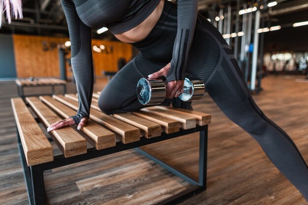 Coole gesunde Fitnessfrau in trendiger Sportbekleidung, die mit Hanteln auf der Bank im Fitnessstudio trainiert und trainiert