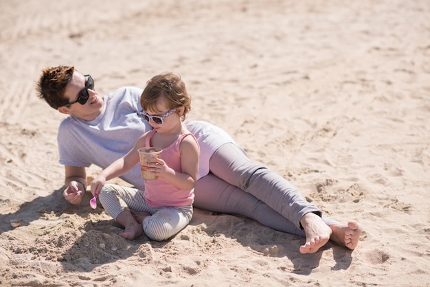 coole junge mutter und ihr kleines mädchen spielen mit einem eimer voller sand mit sonnenbrille und entspannen sich an einem sonnigen tag am strand