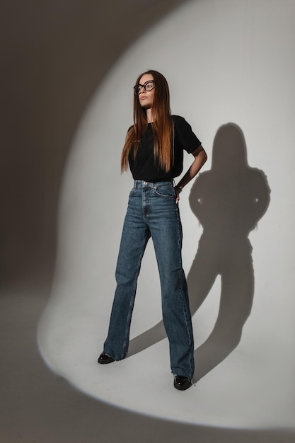 Coole junge stilvolle Frau in Mode schwarzes T-Shirt mit weit ausgestellten Jeans posiert im Studio mit hartem Licht Glamour hübsches Mädchen