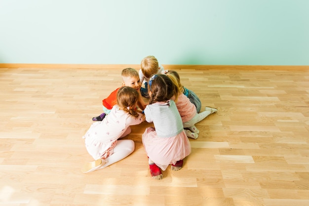 Coole Kinder im Vorschulalter sitzen zusammen in einem kleinen Haufen, verstecken etwas und halten sich an den Händen. Kinder zeigen perfekte Teamarbeit und spielen auf einem Boden in der Kindertagesstätte. Ansicht von oben