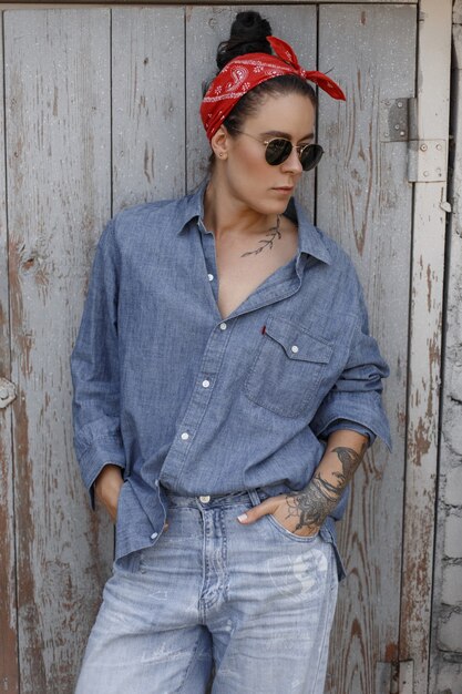 Coole modische Hipster-Frau mit Sonnenbrille mit einem Tattoo auf Händen in einem Jeanshemd und Vintage-Jeans mit einem roten Kopftuch steht in der Nähe einer Holzwand