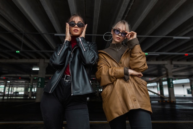 Coole nette Models Freundinnen in modischer Sonnenbrille mit stylischer Lederjacke und Pullover posiert auf einem Parkplatz in der Stadt