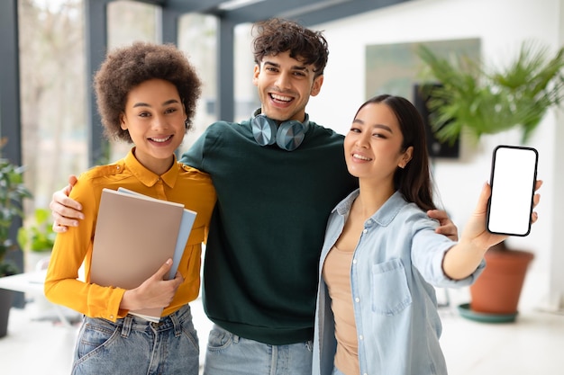 Coole pädagogische Anzeige, glückliche, vielfältige Studenten, die sich umarmen und eine asiatische Dame, die einen leeren Bildschirm auf ihrem Handy zeigt