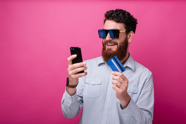 Cooler bärtiger Mann, der sein Telefon betrachtet, das seine Kreditkarte hält, die Sonnenbrille trägt. Internet-Banking oder Online-Shopping-Konzept.