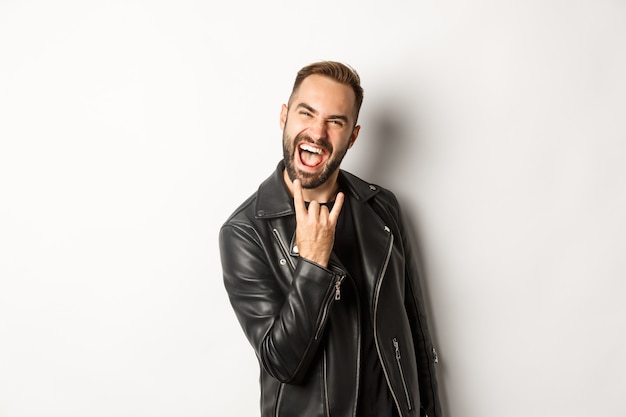 Cooler erwachsener Mann in schwarzer Lederjacke, Rock auf Geste und Zunge zeigend, Musikfestival genießend, auf weißem Hintergrund stehend.