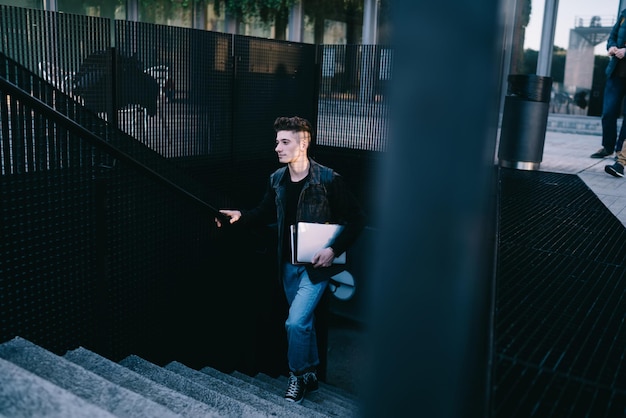 Cooler Hipster-Mann im lässigen Outfit mit Laptop-Gerät, während er die Treppe auf der Stadtstraße hinaufgeht