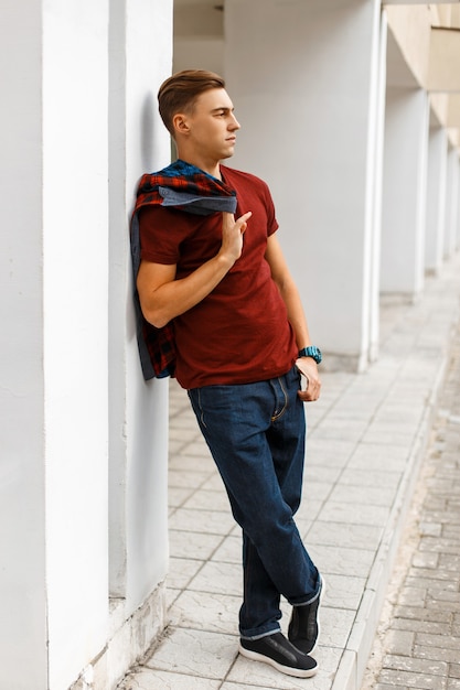 Cooler hübscher junger mann im trendigen roten t-shirt in einem modischen karierten hemd in den blauen jeans in den turnschuhen, die auf der straße nahe einem weißen weinlesegebäude aufwerfen