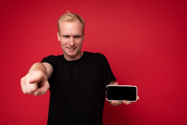 Cooler hübscher positiver blonder junger Mann, der ein schwarzes T-Shirt trägt, das isoliert auf rotem Hintergrund steht