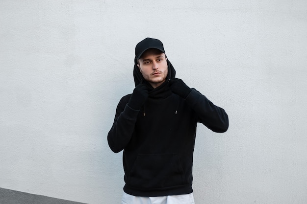 Cooler junger gutaussehender Hipster-Typ in einer stylischen schwarzen Mütze mit einem modischen schwarzen Hoodie steht in der Nähe einer weißen Wand auf der Straße