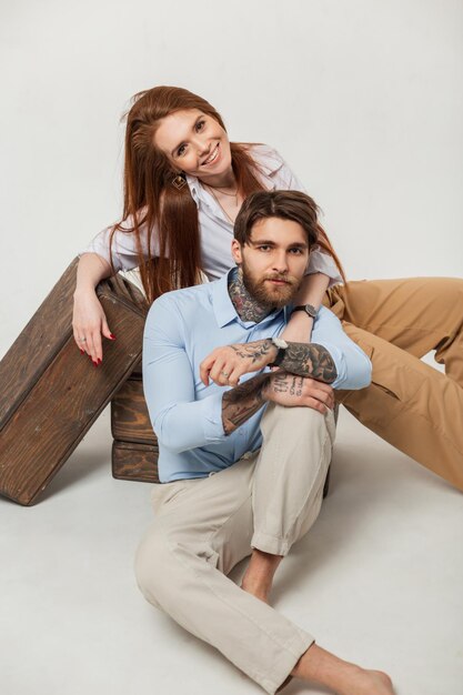 Cooler, stylischer, gutaussehender Hipster-Mann mit Bart und Tätowierungen und schöne, glückliche rothaarige Model-Mädchen sitzen auf weißem Hintergrund Stil Modepaar