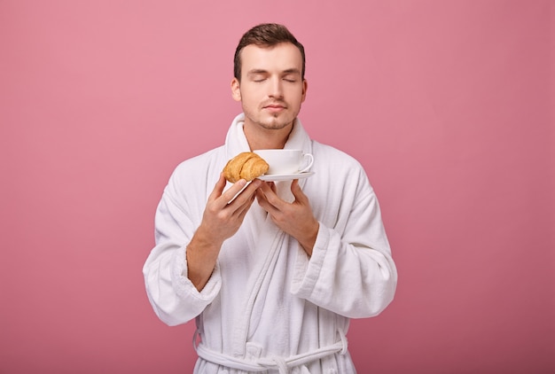 Cooler Typ in Weiß mit duftendem Croissant in einer Hand und einer weißen Tasse und Untertasse in der zweiten