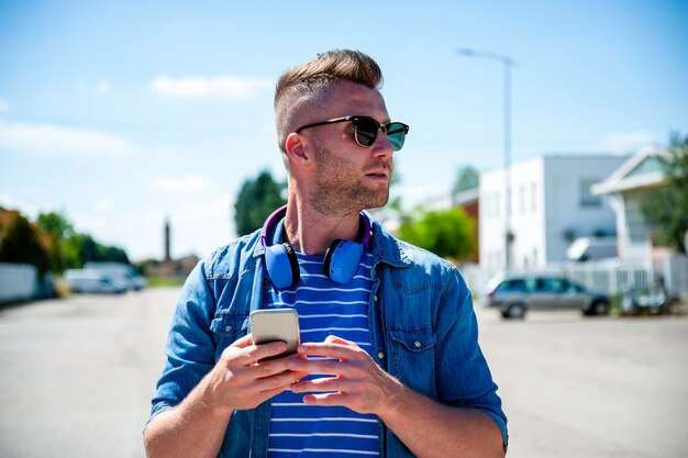 Cooler Typ mit drahtlosem Kopfhörer, der im Freien steht, während er auf dem Smartphone tippt Junger zeitgenössischer Mann in Freizeitkleidung, der sich an sonnigen Tagen auf der Straße amüsiert Urbanes Lifestyle-Konzept