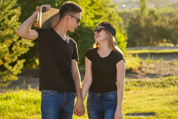 Cooler Typ und ein hübsches Mädchen mit Sonnenbrille, Jeans und einem schwarzen T-Shirt, das Händchen haltend im Sommerpark Skateboard fährt.