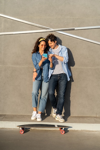 Cooles junges Paar, das draußen auf dem Smartphone-Bildschirm schaut, umarmt den Stand am Longboard-Skate-glückliches Lächeln