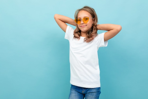 Cooles kaukasisches Teenager-Mädchen im weißen T-Shirt, gelbe Brille lächelt und zeigt sich isoliert auf blauem Hintergrund