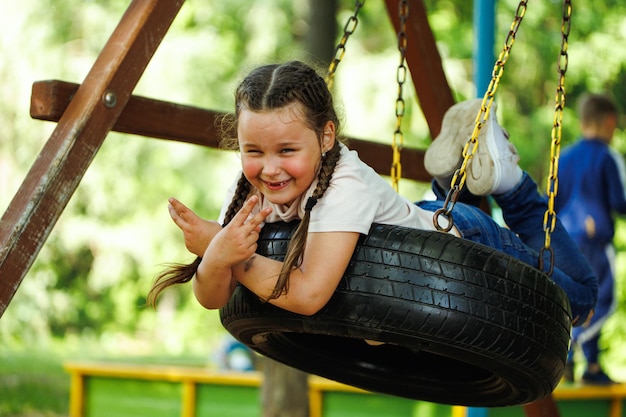 Cooles kleines Mädchen, das auf dem Gummirad auf dem Spielplatz im grünen Park in der Nähe schwingt Sommerferien im touristischen Zentrum des Lagers Gehen und Spielen im Freien, sportliche Aktivität und gesunde Lebensweise Erholung