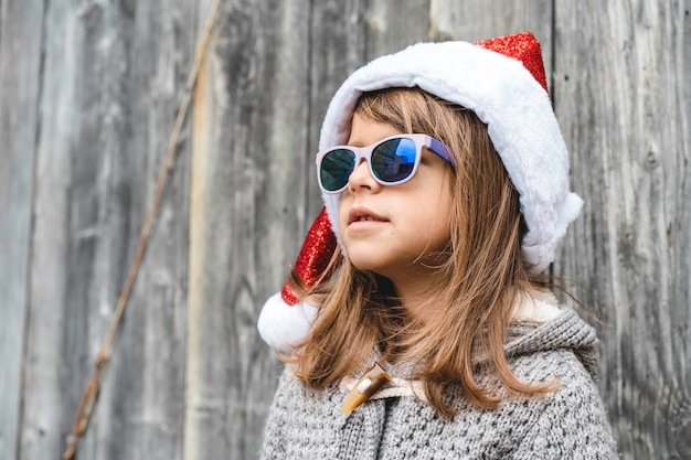 Cooles kleines Mädchen mit Weihnachtsmann-Hut und Sonnenbrille, das in der Weihnachtszeit vor einem hölzernen Grunge-Hintergrund im Freien steht Happy Xmas New Year Winter-Outfit-Konzept und Vintage-Stil