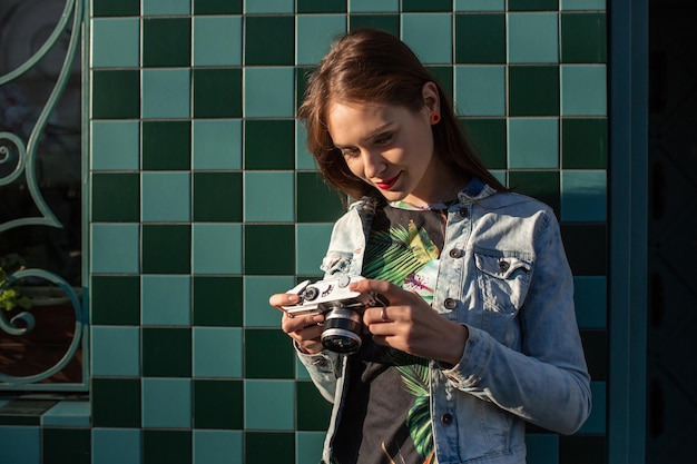 Cooles Mädchenmodell mit Retro-Filmkamera, das eine Jeansjacke trägt, dunkles Haar im Freien über der Stadtmauer in einem Käfighintergrund. Spaß in der Stadt mit der Kamera, Reisefoto des Fotografen.