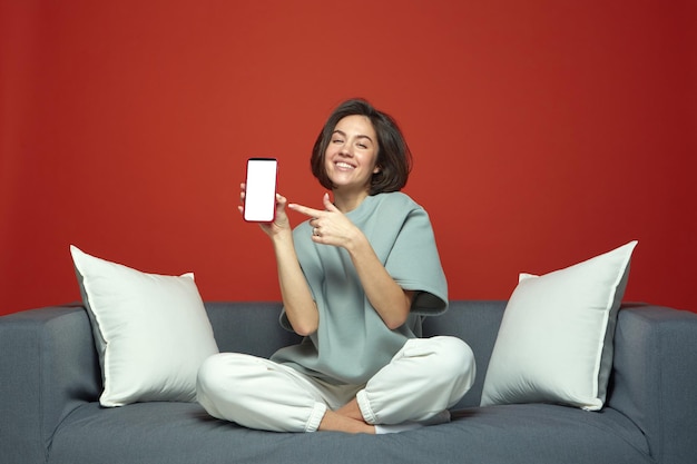 Cooles modernes junges Mädchen zeigt Telefon mit leerem Mockup-Bildschirm, der auf dem Sofa sitzt, neu unterhaltsam oder