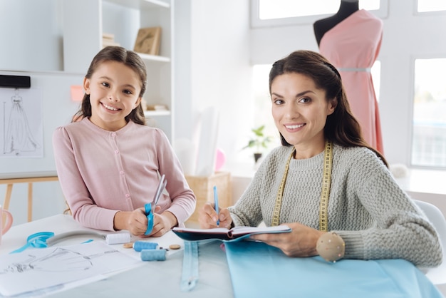 Cooperacion placentera. Diseñadora profesional creativa sentada en la mesa y sonriendo mientras trabaja junto con su hija