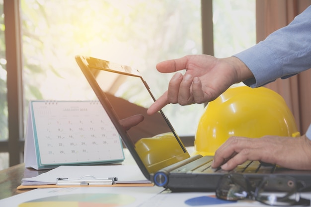 Coordenador do arquiteto que usa o portátil para trabalhar com o capacete amarelo na mesa.