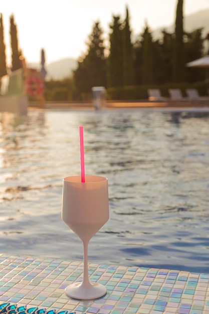 Una copa blanca con una bebida y una pajita rosa se encuentra al borde de la piscina en los rayos