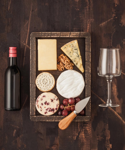 Foto copa y botella de vino tinto con selección de varios quesos en caja de madera y uvas sobre fondo de madera blue stilton red leicester y queso brie y cuchillo