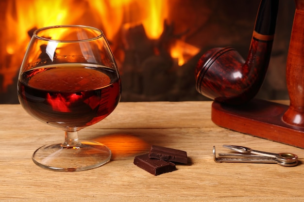 Una copa de brandy, chocolate y pipa de tabaco en la mesa de roble en el fondo de una chimenea encendida