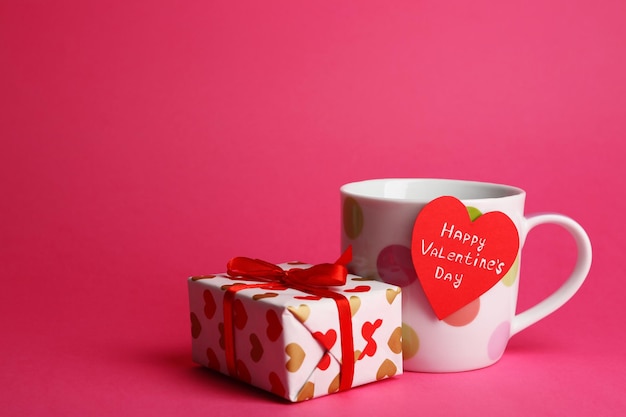 Copa con tarjeta de felicitación y caja de regalo para el Día de San Valentín sobre fondo rosa