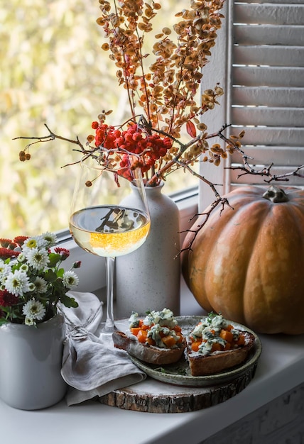 Copa de vino blanco y calabaza gorgonzola bruschetta en el alféizar de la ventana Bodegón de otoño con crisantemos calabaza en el fondo de la ventana Humor de otoño