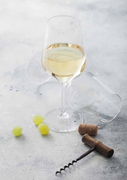 Copa de vino blanco casero con corchos, sacacorchos y uvas sobre fondo de piedra clara con vaso vacío.