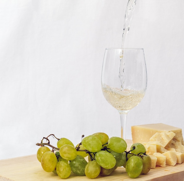 Una copa de vino blanco italiano servido con parmesano y uvas sobre una tabla de cortar de madera con un fondo blanco.